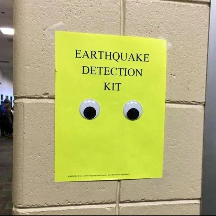 Yellow piece of paper taped to a brick wall. It says "Earthquake detection kit" and has a pair of googly eyes beneath that.