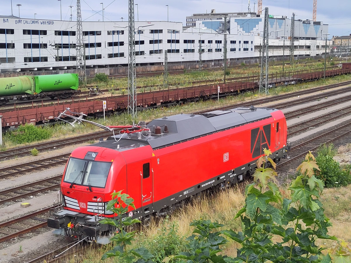 An einem Gleis ohne Oberleitung im Bf Bamberg (NBA) steht eine Vectron Dual Mode in roter DB-Lackierung, deren Stromabnehmer unnatürlich nach vorn abgeknickt ist. Im Hintergrund weitere Gleise mit Güterwagen und Oberleitungen, ganz hinten das seit sechs Jahren leerstehende Einkaufszentrum Atrium.